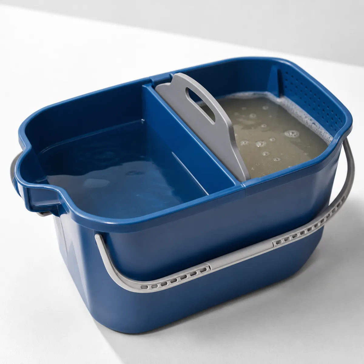 Cutaway of a dual-tank mop bucket showing a clean water compartment and a separate dirty water compartment with a built-in wringer, splash guard and spout